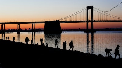 People gather to watch the sun set behind the Throgs Neck Bridge at LIttle Bay Park Wednesday, April 8, 2020, in the Queens borough of New York. While New York Gov. Andrew Cuomo said New York could be reaching a 'plateau' in hospitalizations, he warned that gains are dependent on people continuing to practice social distancing.